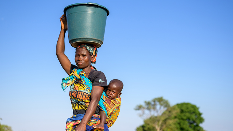 A mother in rural Malawi carries a large bucket of water on her head, while her young baby is swaddled on her back.