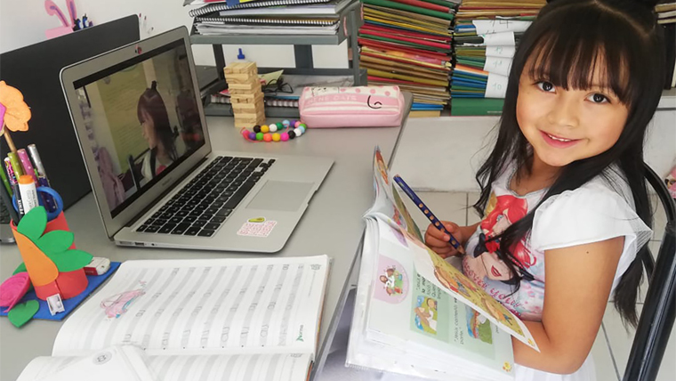 A young girl sits at a table with laptop and books in front of her.
